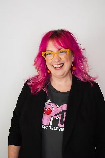 Kristin McCabe smiling, with bright pink hair and yellow cat-eye glasses